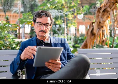 Smart elderly male remote worker in formal wear and trendy eyeglasses sitting with crossed legs on bench and working with digital tablet at park Stock Photo