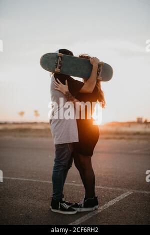 Side view of romantic teenage couple embracing and covering heads with skateboard while kissing in back lit of sunset on rural road Stock Photo