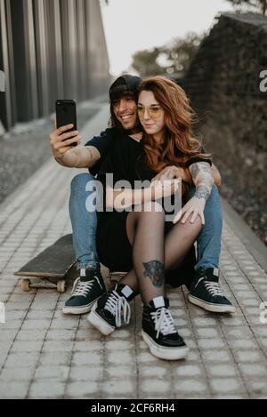 Cool trendy couple sitting on road with skateboard and taking selfie together with mobile phone in city Stock Photo