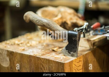 Shabby axe with wooden handle sticking out of large square wooden table with sawdust on background of carpentry tools and piece of wooden raw material in workshop Stock Photo