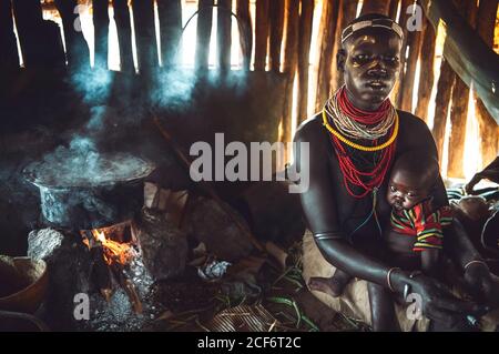 Omo Valley, Ethiopia - November 08, 2018: Woman from Karo tribe embracing cute baby while sitting near heart and pot in hut of village in Ethiopia Stock Photo