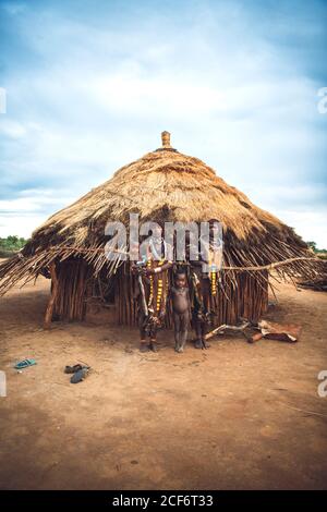 Omo Valley, Ethiopia - November 08, 2018: Two women of Hamer tribe standing with babies against village hut. Omo valley, Ethiopia Stock Photo