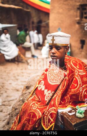 Lalibela, Ethiopia - November 04, 2018: Young black man wearing cloak and headgear of Orthodox priest sitting outdoors in rock-hewn church of Lalibela, Ethiopia Stock Photo