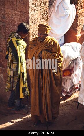 Lalibela, Ethiopia - November 04, 2018: Senior ethnic man in long gown standing on rock of ancient monolithic church with prayers, Ethiopia Stock Photo