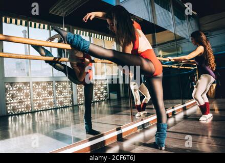 Confident young sportive women in bodysuits practicing in ballet studio stretching with barre. Stock Photo