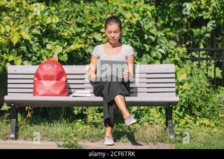 University student watching a live stream video of an online class remote learning during covid. Asian woman using laptop to study in city park bench Stock Photo