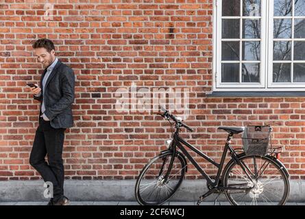 Young businessman going to work on bike commute using his mobile cellphone against city brick wall background. Happy business man bicycle commuter Stock Photo