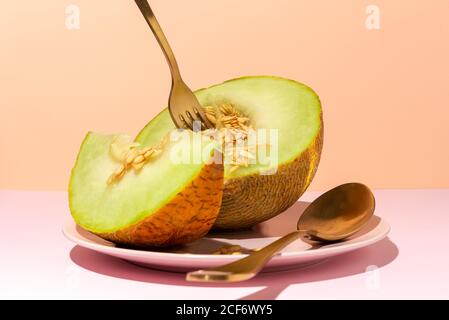 Cut ripe appetizing sweet pitted melon on plate with spoon and fork in pink table Stock Photo