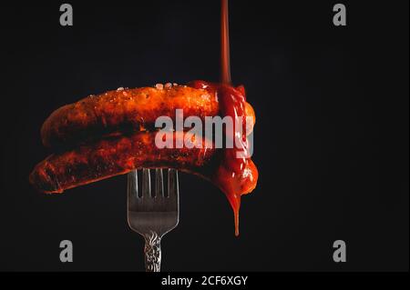 ketchup falling on delicious grilled sausages pricked in fork in a black background Stock Photo