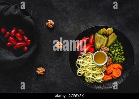 Zucchini raw vegan pasta with peas, cherry tomatoes, avocado; carrot; beet; Nuts, olive oil on bowl. On dark table from above. Vegetarian healthy food. Flat lay Stock Photo