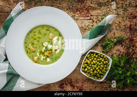 From above mashed vegetable creamy soup with small crackers and green peas and parsley in white bowl on wooden background Stock Photo