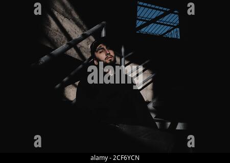 Young bearded man in stylish urban outfit looking at camera while leaning on railing inside shabby abandoned building Stock Photo
