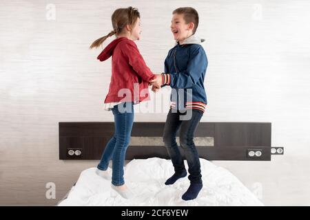 Side view of playful boy and girl holding hands and jumping on soft bed while having fun during weekend Stock Photo