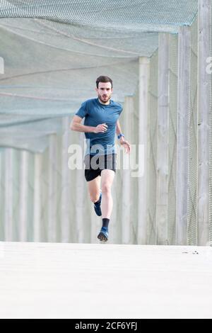 Highly motivated bearded male athlete in blue t shirt and shorts running outdoors under cover looking at camera Stock Photo