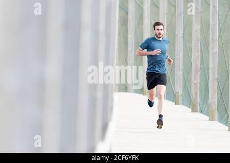 Highly motivated bearded male athlete in blue t shirt and shorts running outdoors under cover looking at camera Stock Photo