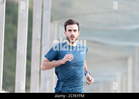 Highly motivated bearded male athlete in blue t shirt and shorts running outdoors under cover looking at camera Stock Photo