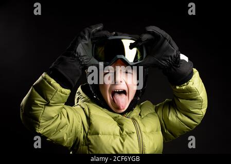 Focused little sportive kid in winter clothes putting on ski goggle on black background in studio looking at camera Stock Photo