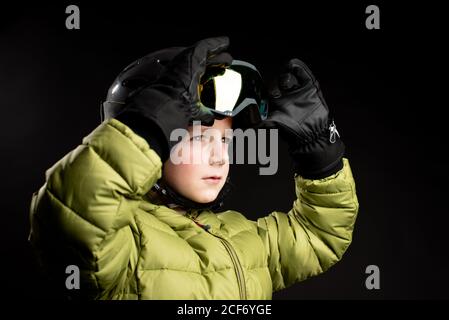 Focused little sportive kid in winter clothes putting on ski goggle on black background in studio Stock Photo