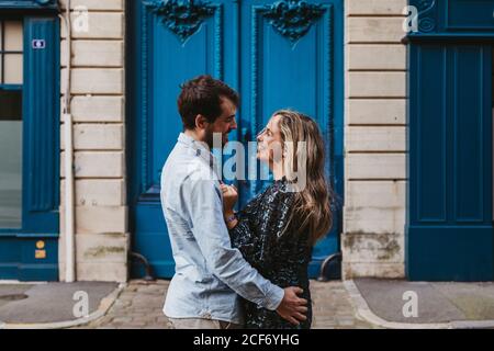 Side view of happy young couple in casual clothes hugging looking at each other while standing against aged stone building with blue doors on city street Stock Photo