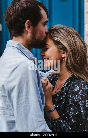 Side view of happy young couple in casual clothes hugging and kissing while standing against aged stone building with blue doors on city street