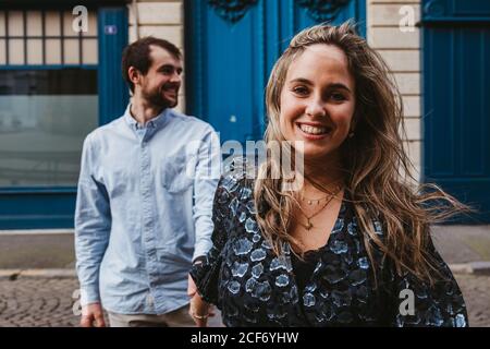 Happy young Woman looking at camera in stylish outfit followed by smiling boyfriend walking on city street with old building in background during romantic holidays in France Stock Photo