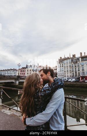 Side view of happy young affectionate couple embracing and kissing while standing on stone embankment near river with old buildings in background in Bayonne city in France Stock Photo