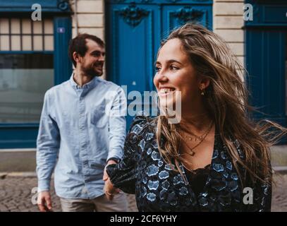 Happy young Woman in stylish outfit followed by smiling boyfriend walking on city street with old building in background during romantic holidays in France Stock Photo