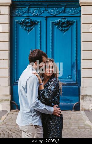 Side view of happy young couple in casual clothes hugging and kissing while standing against aged stone building with blue doors on city street Stock Photo