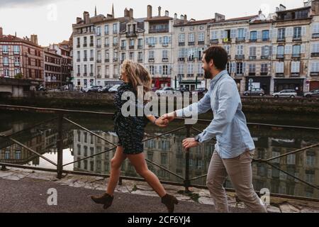 Side view of happy young affectionate couple running with holding hands laughing on stone embankment near river with old buildings in background in Bayonne city in France Stock Photo