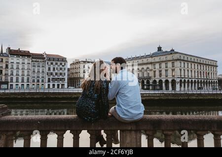 Back view of happy couple sitting embracing each other on old stone fence spending summer evening together in Bayonne Stock Photo