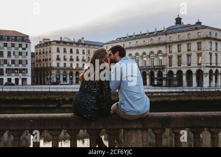 Back view of happy couple sitting kissing each other on old stone fence spending summer evening together in Bayonne Stock Photo