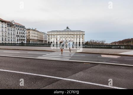 Happy young romantic couple in stylish clothes laughing and holding hands while crossing bridge with historic buildings in background during city tour in Bayonne in France Stock Photo