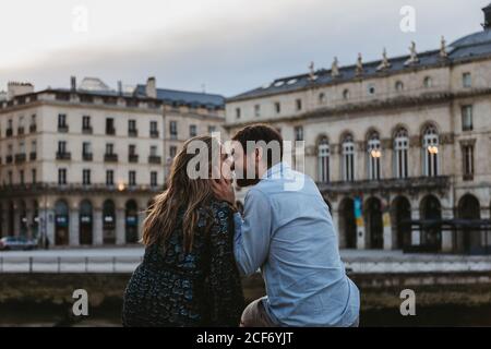 Back view of happy couple sitting kissing each other on old stone fence spending summer evening together in Bayonne Stock Photo