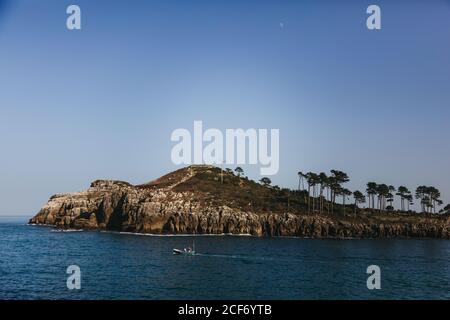 Lonely fishing boat moving on bay with tranquil water against rocky shore with tall evergreen trees at foot of hill under clear blue sky in Spain Stock Photo