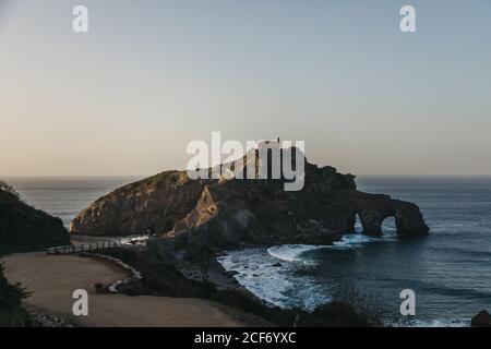 Empty road leading along bridge and ridge to lonely country house on top of rocky hill on island Gaztelugatxe surrounded by calm sea water with white foam waves in sunny evening Stock Photo