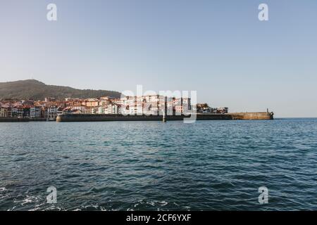 Picturesque scenery with blue water of bay with small waves against waterfront of colorful town at foot of forested hill under cloudless sky in Spain Stock Photo