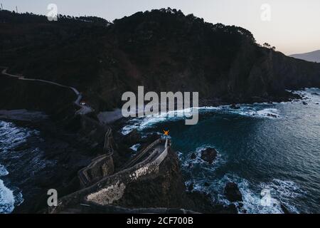 From above back view of person with outstretched arms enjoying freedom standing on old stone bridge against troubled water with foam waves washing rocky shore in Spain Stock Photo