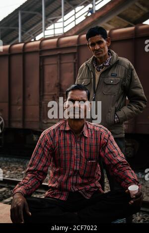 Allahabad city, India - FEBRUARY, 2018: Serious adult Indian man in checked shirt with cup of drink in hand sitting with crossed legs on rails while colleague in jacket standing behind and both looking at camera with freight wagon in background Stock Photo