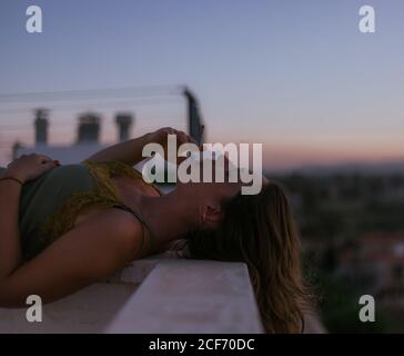 Side view of peaceful lady lying with closed eyes on fence of balcony and smoking cigarette with sunset on blurred background Stock Photo