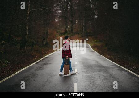 Side view of young stylish man in casual wear walking on asphalt road with skateboard in hand on autumn day Stock Photo