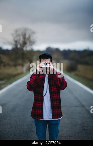unrecognizable man in stylish casual outfit holding photo camera in front of face while standing on road in countryside Stock Photo