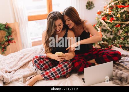 Two women friends enjoying in winter holidays at home and looking something on mobile phone near christmas tree in cozy interior. Interior with christmas decorations. Stock Photo