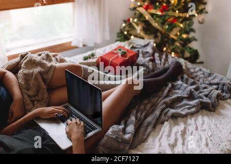 Two friends with knee high socks in winter holidays at home writing something on laptop near christmas tree in cozy interior with christmas presents near christmas tree.
