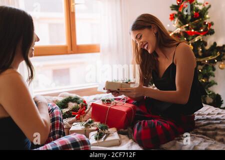 Portraits of two friends opening gifts in cozy bed near christmas tree. Closeup legs of women at home. Stock Photo