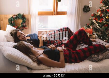 Two fun beautiful friends smiling each other in bed at home near christmas tree in cozy interior. Interior with christmas decorations. Stock Photo