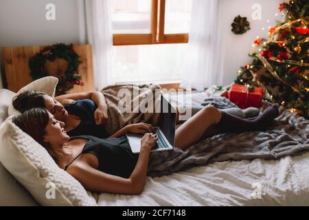 Two women friends in winter holidays at home looking something on laptop near christmas tree in cozy interior. Interior with christmas decorations. Stock Photo