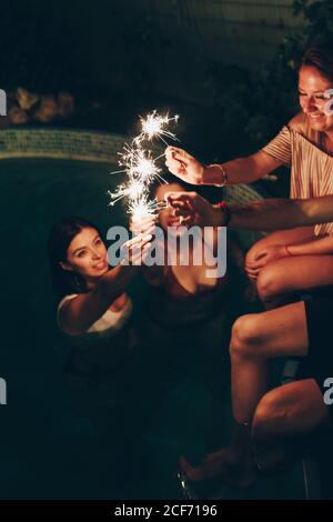 Group of young people smiling and burning sparklers while having party near pool at night Stock Photo