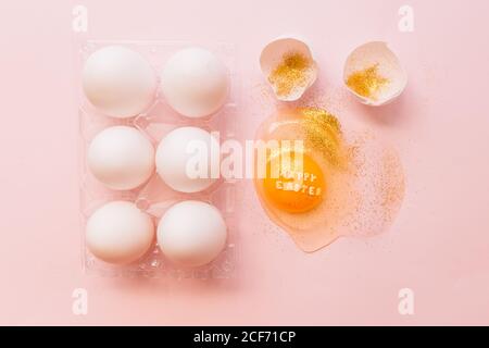 Top view of broken glittered egg with text Happy Easter and raw eggs in plastic box on pink background Stock Photo