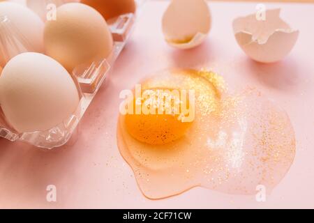 Top view of broken glittered egg with text Happy Easter and raw eggs in plastic box on pink background Stock Photo