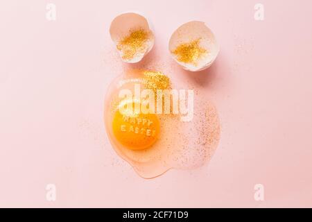 From above raw broken egg with text Happy Easter on yellow yolk covered in glitter and broken eggshells on pink background Stock Photo